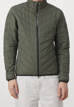 Homme portant une veste matelassée ajustée de couleur olive avec une fermeture éclair à l'avant et un col haut, associée à un pantalon clair.