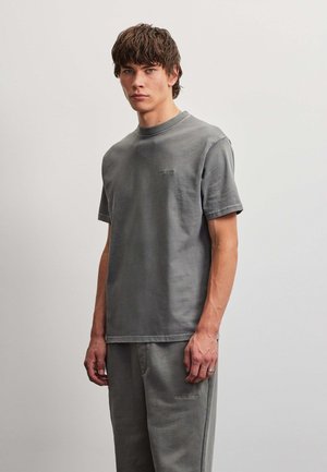 Jonge man met kort krullend haar draagt een bijpassend gewassen grijs T-shirt en broek en staat tegen een effen lichte achtergrond.