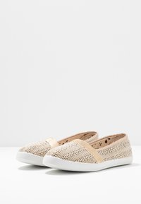 Chaussures slip-on tissées en beige et or, avec un dessus texturé, une bande d'accent dorée et une semelle en caoutchouc blanche. Les imprimés intérieurs sont noirs sur beige.