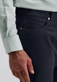 Nahaufnahme einer Hand eines Mannes in der Nähe der Vordertasche von dunklen Cordhosen und dem Ärmel eines weißen Langarmhemdes.