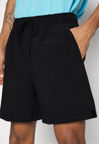 Homme portant un short noir Nike avec une taille à cordon de serrage et des poches latérales, main posée dans la poche droite, chemise bleu clair visible.