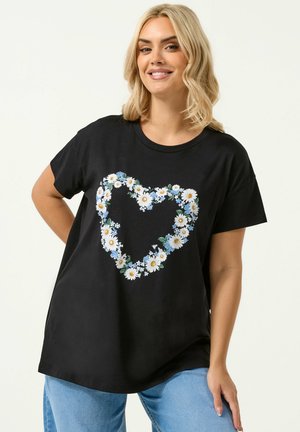 Blondynka uśmiechająca się, ubrana w czarny t-shirt z sercem zrobionym z białych stokrotek i niebieskich kwiatów, do tego jasnoniebieskie dżinsy.