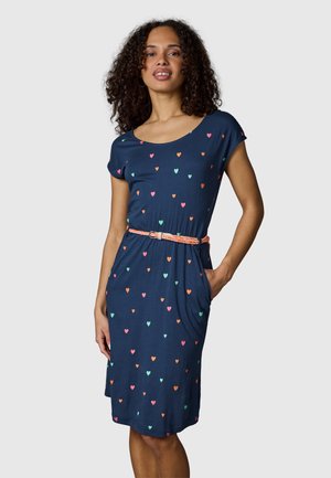 Vrouw in marineblauwe knielange jurk met meerkleurige hartjesprints, oranje gevlochten riem, één hand in de zak, staand tegen een effen achtergrond.