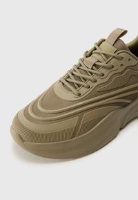 Chaussure de sport vert olive avec un dessus texturé, un design épuré, une semelle en caoutchouc proéminente et un système de laçage avec une languette au talon.