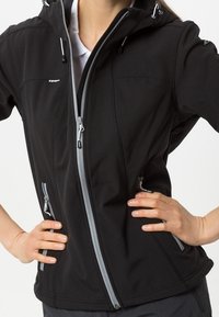 Person iført en sort lynlåsjakke med grå lynlåsdetaljer, hænderne hvilende på hofterne, stående foran en ensfarvet baggrund.