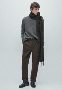 Maglione grigio lavorato a maglia con collo alto, abbinato a pantaloni plissettati marrone scuro e una lunga sciarpa nera. Scarpe nere completano l'outfit.