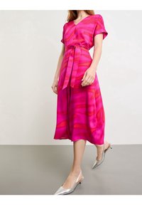 Różowa midi sukienka z krótkimi rękawami, z paskiem w talii, z abstrakcyjnymi jaśniejszymi różowymi i czerwonymi wzorami. W połączeniu z srebrnymi butami na szpilkach.