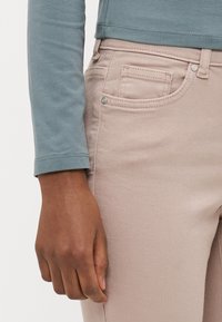 Top de manga larga en color verde azulado con escote redondeado, combinado con jeans de mezclilla rosa claro. Bolsillo frontal visible y detalle de botón metálico en los jeans.