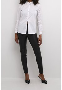 Camisa blanca de botones con cuello, combinada con pantalones de cuero negros ajustados y tacones negros de punta con acentos decorativos de hardware.