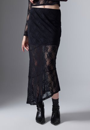 Woman wearing black lace midi skirt over shorter lining and black pointed leather ankle boots against gray background.