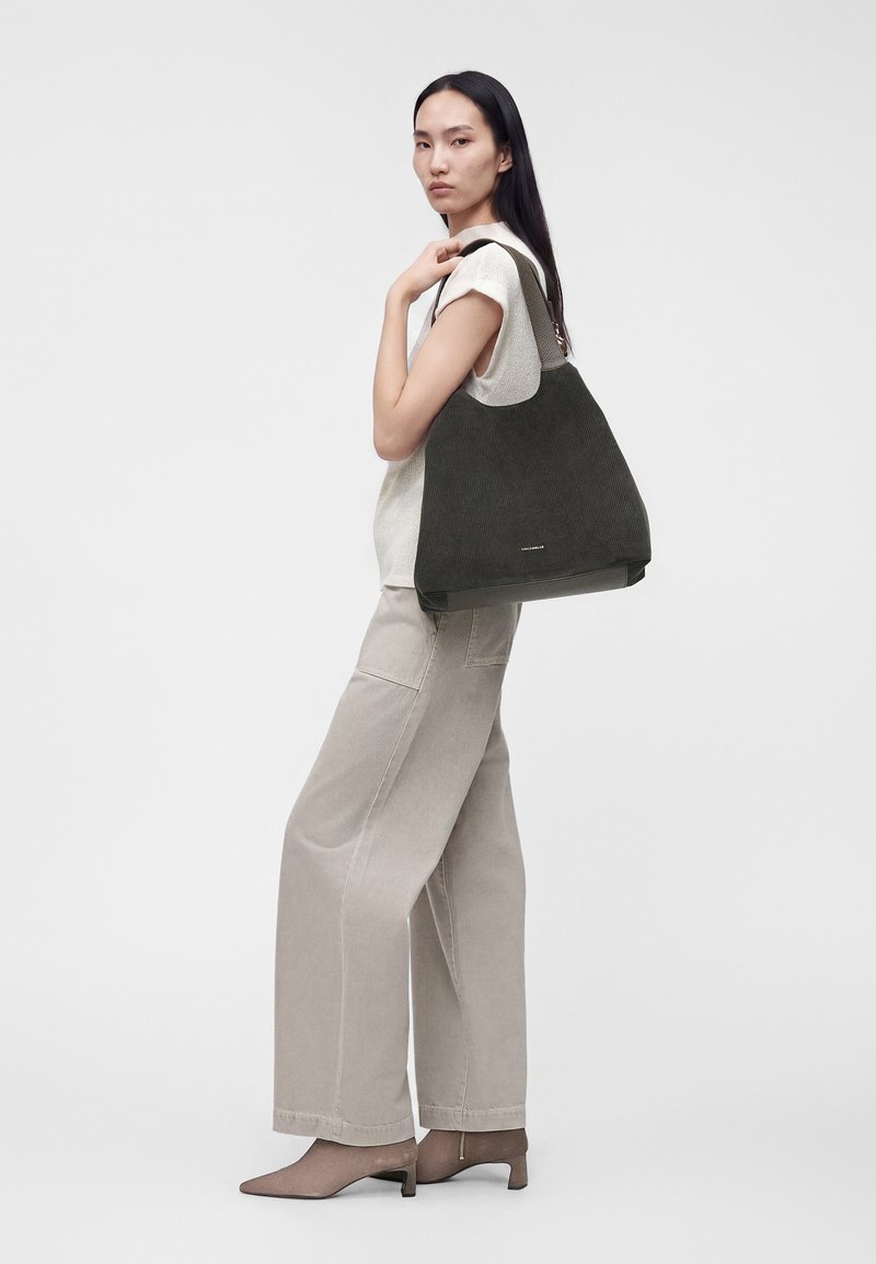 Femme portant un haut blanc sans manches et un pantalon large beige, portant un grand sac cabas vert foncé sur l'épaule sur fond blanc.