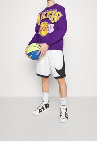 Sweat-shirt violet avec le logo "Los Angeles Lakers", shorts blancs avec accents noirs, et baskets noir et blanc. Tenant un ballon de basket multicolore.