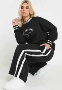 Ensemble de sport noir comprenant un sweat-shirt à col bateau avec des lettres blanches et des poignets rayés, associé à un pantalon large avec des rayures blanches sur les côtés.