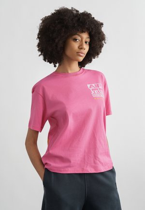 Νεαρή γυναίκα με σγουρά μαλλιά, που φοράει ροζ μπλουζάκι Air Jordan World Tour και σκούρο παντελόνι, στέκεται με τα χέρια στις τσέπες μπροστά σε απλό φόντο.