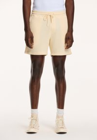 Waffle-knit beige shorts with an elastic waistband and drawstring, featuring side pockets and a relaxed fit. Worn with beige high-top sneakers.