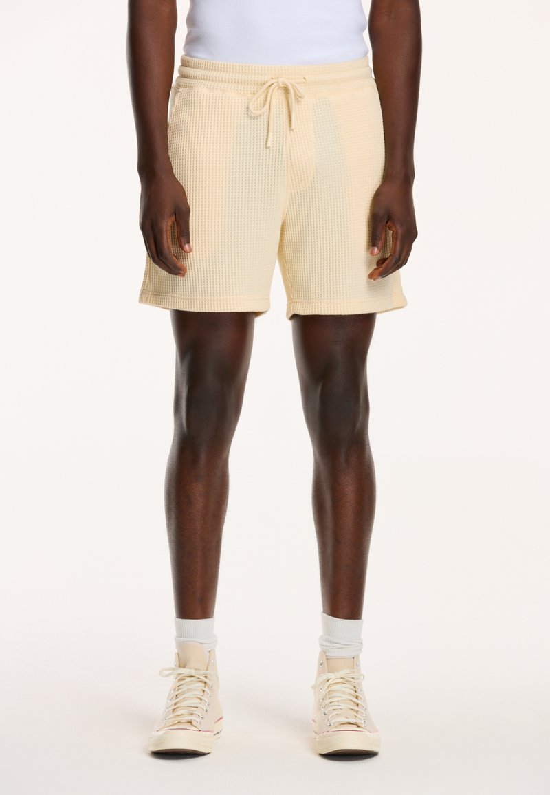Waffle-knit beige shorts with an elastic waistband and drawstring, featuring side pockets and a relaxed fit. Worn with beige high-top sneakers.