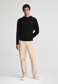 Zwarte kabelgebreide trui met een ronde hals, gecombineerd met beige rechttoe-rechtaan broek en lichte sneakers. Model staat tegen een neutrale achtergrond.