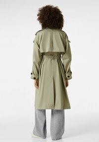 Khaki trenčkot s páskem v pase, ramenními epaulety a velkými manžetami. Látka je hladká a design má středně dlouhý střih.