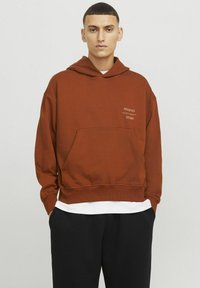 Bruine hoodie met een frontzak, met de teksten "ORIGINALS" en "STUDIO". Het heeft een relaxed fit en is gelaagd over een wit shirt.