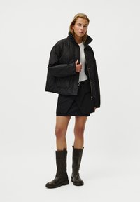 Black quilted jacket with a high collar, paired with a white top and black skirt. Worn with tall brown leather boots featuring a chunky sole.