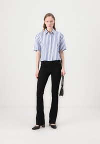 Afgekorte, button-up shirt met blauw en witte strepen, gecombineerd met smalle zwarte broeken en zwarte hakken; model houdt een kleine zwarte tas vast.