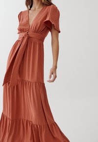 Rostfärgad maxiklänning med v-ringning, korta utsvängda ärmar, detaljer med knytband i midjan och lager av volanger vid fållen. Strukturerad tyg.