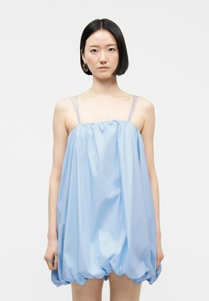 Femme aux cheveux noirs et raides portant une robe ample bleu clair sans manches à fines bretelles, debout devant un fond blanc uni.