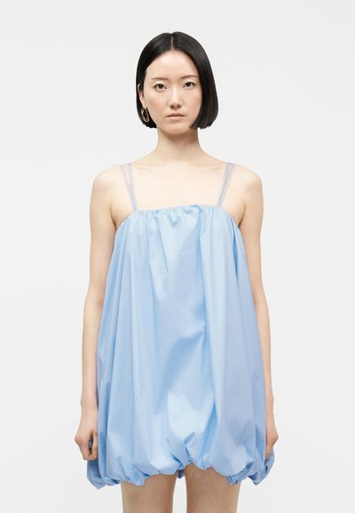 Femme aux cheveux noirs et raides portant une robe ample bleu clair sans manches à fines bretelles, debout devant un fond blanc uni.