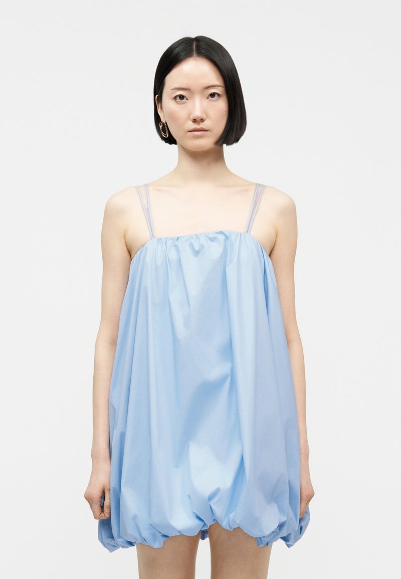 Femme aux cheveux noirs et raides portant une robe ample bleu clair sans manches à fines bretelles, debout devant un fond blanc uni.