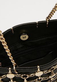Bolso negro con asas de cadena dorada, cierre de botón magnético y un bolsillo interior con cremallera visible en el interior.