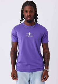 Camiseta corta de manga en color púrpura hecha de algodón, con un logo blanco "PXP" y un diseño de estrella en el pecho. Combinada con pantalones vaqueros de color azul claro.