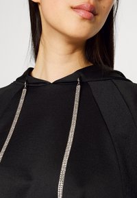 Czarna bluza z kapturem o fakturowanym wykończeniu, wyposażona w srebrne metalowe elementy i ściągacz ze sznurkiem umożliwiający regulację dopasowania. Zbliżenie.