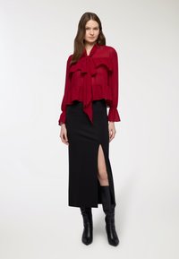 Blusa rossa trasparente con dettagli a volant e colletto a fiocco, abbinata a una gonna nera a maxi aderente con spacco laterale. Stivali neri al ginocchio.