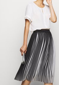 Femme portant une blouse blanche et une jupe plissée rayée verticale noir et blanc, tenant une pochette en argent.
