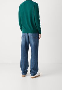 Maglione verde lavorato a maglia con orlo a coste, abbinato a jeans in denim blu larghi e sneaker bianche. Vista da dietro, che mette in mostra le texture del capo.