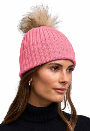 Kobieta nosząca różową prążkowaną dzianinową czapkę z puszystym pomponem na czubku i srebrne kolczyki koła, patrząca w prawo, na białym tle.