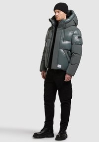 Szara pikowana kurtka z wysokim kołnierzem, dużym kapturem i zamkiem z przodu. Noszona na czarnej bluzce, zestawiona z czarnymi spodniami typu cargo i butami.