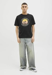 Camiseta de algodón negra con un gráfico circular que presenta palmeras y el texto "Palm Spring" en naranja y amarillo. Combinada con pantalones de mezclilla claros.