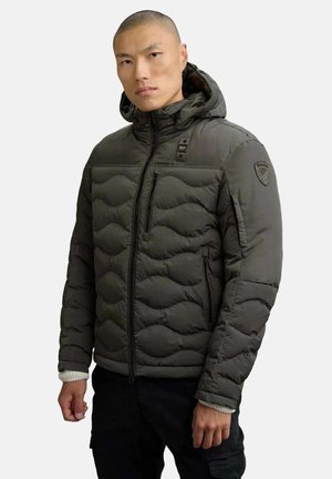 Uomo che indossa un giubbotto imbottito verde oliva con cappuccio, dotato di tasche con zip sul davanti e sul petto, in piedi con un'espressione neutra contro uno sfondo semplice.