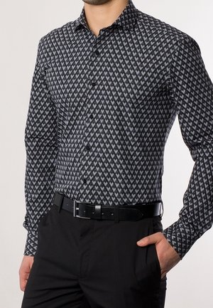 Chemise à manches longues noire et gris à motifs géométriques, boutonnée, portée avec un pantalon noir et une ceinture noire, une main dans la poche.