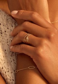 Trois bagues en or empilées sur une main au toucher lisse, reposant sur une surface en tricot beige. Un bracelet en or simple est visible à proximité.