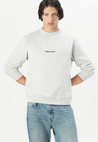 Lichtgrijze sweatshirt met geribbelde manchetten en onderrand. "Original Studio" logo in het zwart geborduurd op de borst. Gemaakt in combinatie met blauwe spijkerbroek.