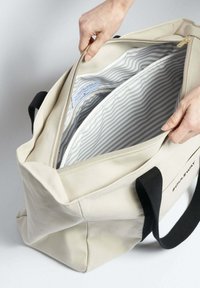 Béžová plátěná taška s černými uchy, vybavená zipovým uzávěrem a pruhovanou šedou podšívkou uvnitř. Viditelné výrazné švy.