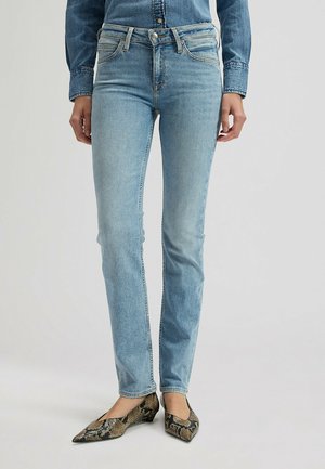 Persoon draagt lichtblauwe slim-fit jeans, een spijkershirt in de broek gestopt en puntige flats met slangenpatroon, staand op een effen lichtgrijze achtergrond.