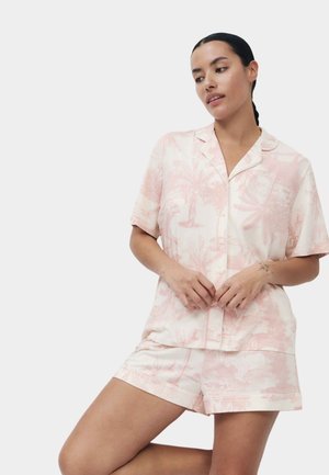 Femme aux cheveux foncés en tresse basse porte une chemise à manches courtes boutonnée à motifs rose pâle et un short assorti sur un fond uni.