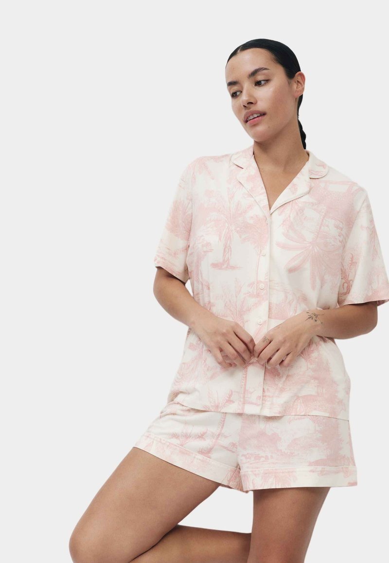Vrouw met donker haar in een lage vlecht draagt een lichtroze korte mouwen blouse met patroon en knopen en bijpassende korte broek tegen een effen achtergrond.