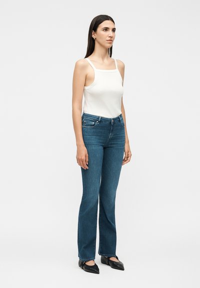 Γυναίκα που στέκεται ελαφρώς γυρισμένη προς τα δεξιά, φορώντας λευκό αμάνικο τοπ, μπλε τζιν και μαύρα μυτερά παπούτσια, με απλό φωτεινό φόντο.