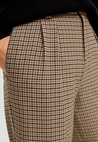 Pantalon à carreaux marron et noir avec plis, coupe ajustée et tissu texturé. Poches visibles et détail à la taille inclus.