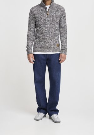 Uomo che indossa un maglione grigio con zip e motivo a disegni, maglietta bianca sotto, jeans blu scuro e scarpe da ginnastica bianche, in piedi su uno sfondo bianco.