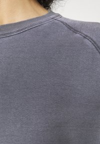 Primer plano de la parte posterior del hombro y el área del cuello de una persona que lleva una camiseta de cuello redondo de color púrpura desvaído con detalles de costura visibles.